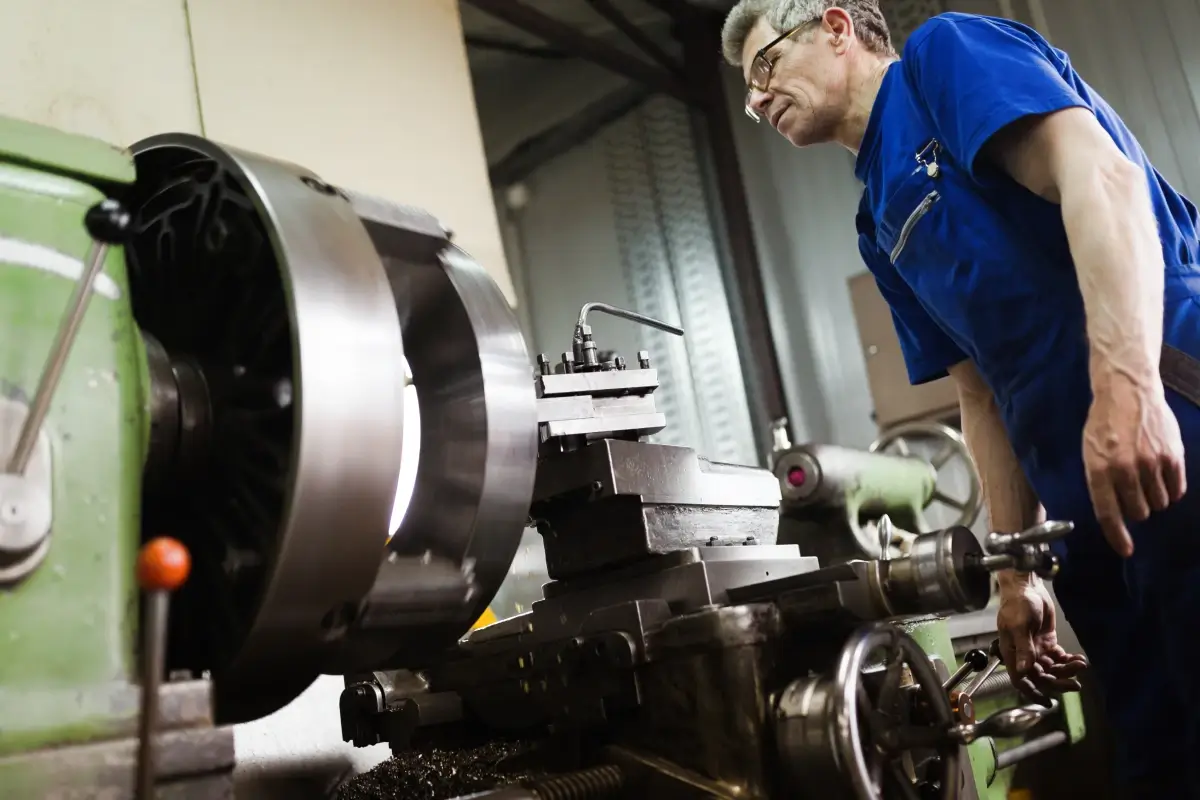 Machinist operating an industrial lathe while inspecting a spinning metal workpiece in a manufacturing shop.