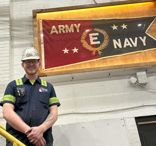 Mason, a level 8 assembler, smiles while wearing a hard hat, standing next to an Army/Navy flag.