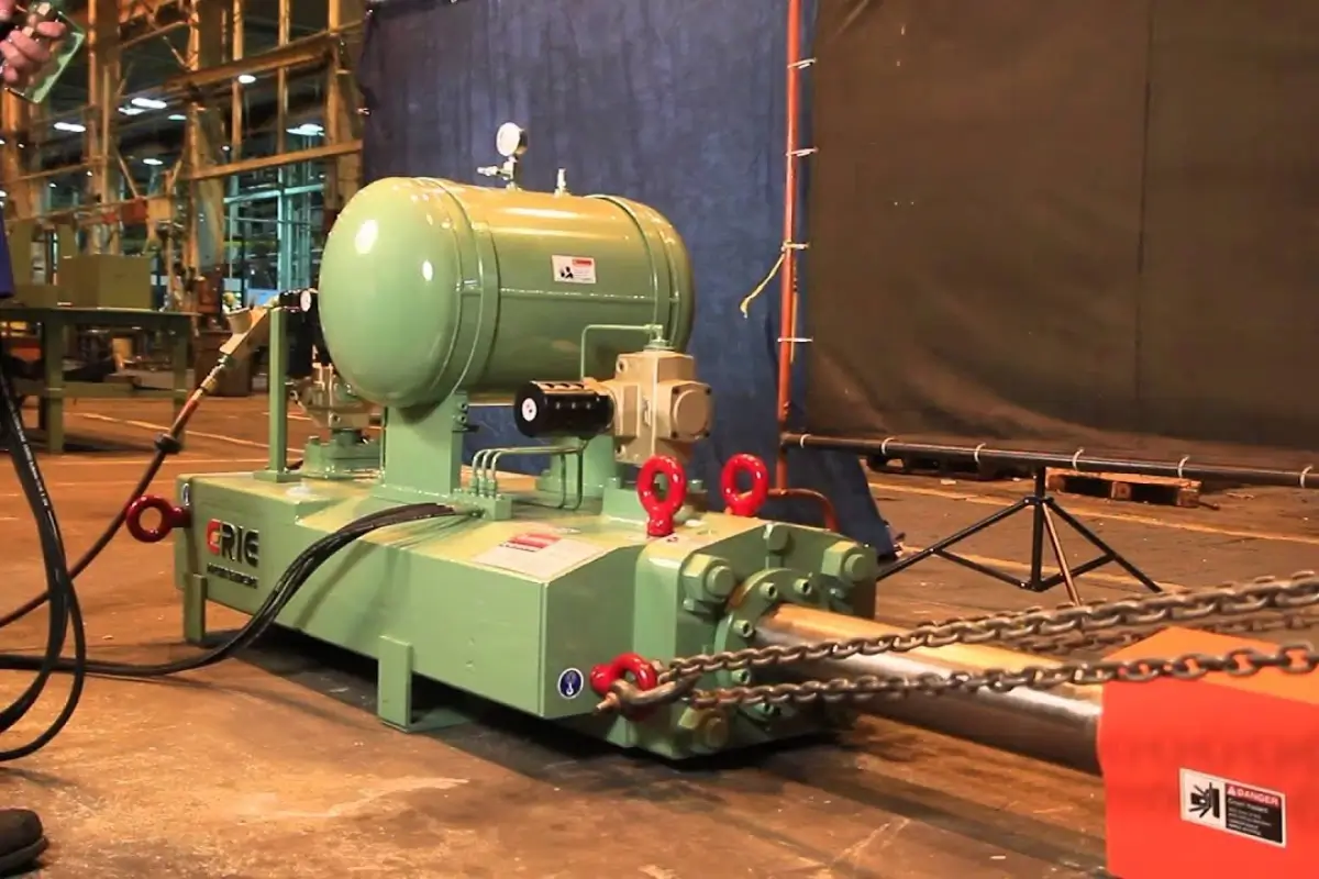 Green pneumatic forging hammer with air pressure tank, gauges, and chained hammer ram on a factory floor.